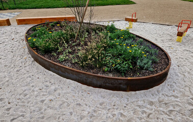 the oval ellipse of the flower bed is lined with a rusty sheet metal border. perennials are blooming. sandpit with park features and children's playground. rounded edge, stepping stones, concrete