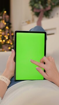 Woman Uses Tablet Computer With Green Mock-up Screen Lying On Sofa In Cozy Room With Christmas Tree And Lights. Girl Surfing Internet and Making Online Shopping