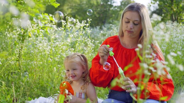 A mother plays with her daughter sitting in the grass in the field blowing soap bubbles. The concept of happiness and children's games with fairy tales of smiles. High quality 4k footage