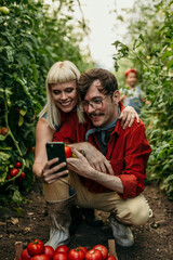 A bonding loving family happily picking fresh, organic tomatoes in their greenhouse and using a phone.