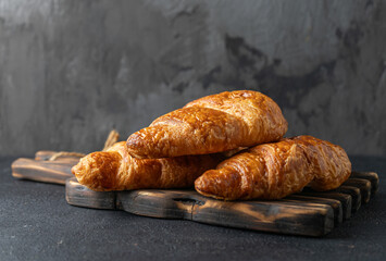 Fresh croissants with a golden brown and crispy crust on a wooden board and against the background of an old dark concrete wall.