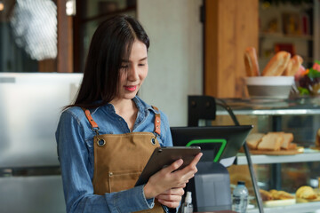 Beautiful young Asian barista holds tablet to show customers menus and smiled standing in front of counter, and next to it was a bakery cabinet with bread, cakes, and products from small coffee shop. - obrazy, fototapety, plakaty
