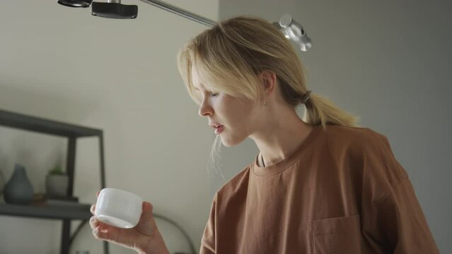 Medium close-up tilting shot of young Caucasian female blogger with blonde ponytail looking at jars and pots of face cream, while taking promotional flatlay photos in home studio