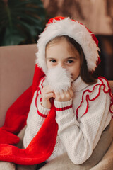 Close-up Caucasian child, lovely baby girl in Santa hat, makes cherished wish and cute presents, surrounded by Christmas decorations. Magic festive atmosphere at home
