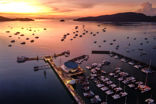 Aerial drone view of Chalong Pier during sunrise in Phuket, Thailand. Many yachts, catamaran and speed boats moored at the pier platform of Ao Chalong Bay, one of centers to travel around Andaman Sea.