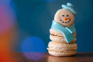 Snowman macaron dessert decorated with hat and scarf against colorful lights and bokeh background. Merry Christmas and Happy New Year festive concept