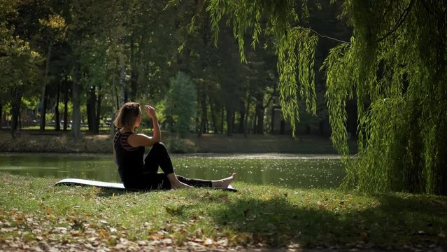 Woman sitting on yoga mat and practicing yoga stretching exercise outdoors in sunny day. Wide footage