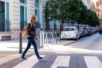Visually impaired woman with white cane in hand walking through pedestrian crossing in the city - obrazy, fototapety, plakaty