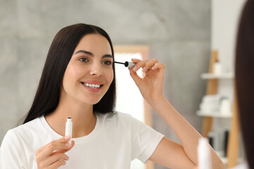 Beautiful woman applying serum onto her eyelashes indoors, reflection in mirror. Cosmetic product