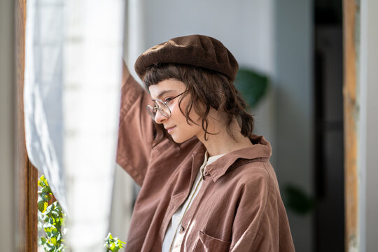 Pensive melancholic teenager in vintage clothes, beret and glasses standing at window ang looking attentively. Teen girl with concentrated glance looks like dreamer, bookworm, brainy diligent student 