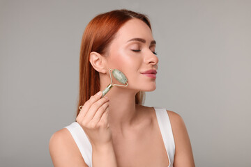 Young woman massaging her face with jade roller on grey background