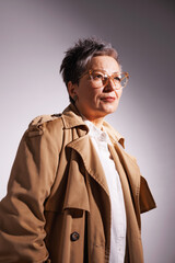 Mature woman in trench coat and eyeglasses looking away isolated on grey