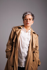 Confident and elegant mature woman in trench coat, white shirt and eyeglasses standing isolated on grey 