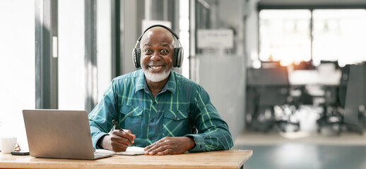 portrait of happy bald and bearded african american senior man wearing headphones, enjoying working at home office.