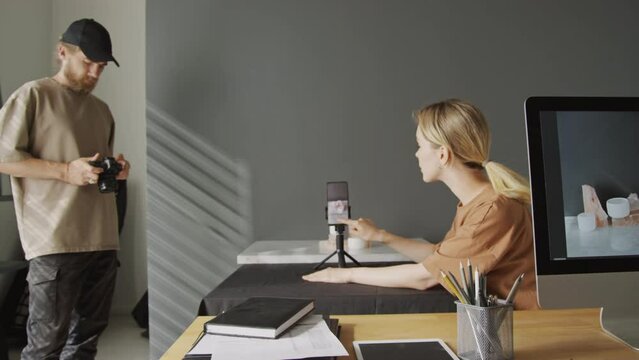 Medium shot of young blonde Caucasian woman sitting at table in studio, and taking photos of commercial product samples on smartphone on tripod, and male colleague with dslr camera watching