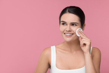 Young woman cleaning her face with cotton pad on pink background. Space for text
