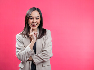 A portrait of an Indonesian Asian woman wearing a cream-colored blazer, posing happily with an index finger on her cheek. Isolated against a magenta background.