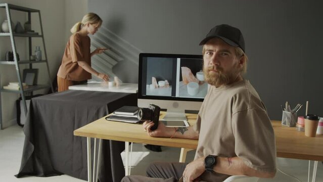 Medium portrait shot of young Caucasian man with red beard, in baseball cap and casual t-shirt sitting at desk in front of computer monitor with product sample photos and looking at camera