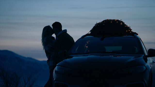 Silhouette of a couple kissing near the car with lit Christmas tree tied to the roof, winter mountains in the background