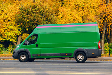 A green van drives along the road next to a city park in autumn. Fall has arrived, trees in...
