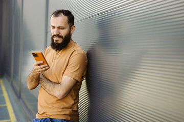 Caucasian bearded guy using smartphone leaning on a urban wall