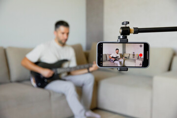 A man records playing guitar on his phone; the mobile phone is mounted on a tripod to record video....