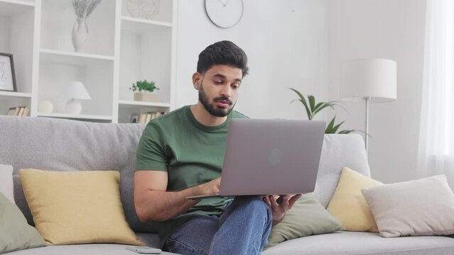 Indian young man shaking his head in disbelief while working on modern laptop at home. Male freelancer looking on screen with disappointment on face while sitting on comfy couch.