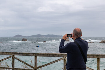 Obraz premium Senior man on his back photographing the sea at sunset with a camera while raining. Summer tourism. Galicia. Active seniors. Copy space.
