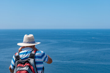 Senior man on his back with white sun hat and backpack photographing the Atlantic ocean. Tourism in...