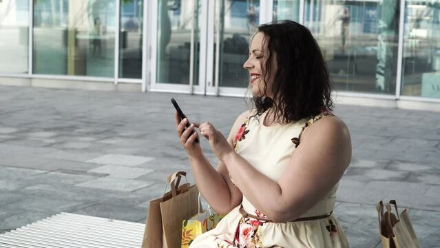 Portrait of 40s plus size woman with sitting on a bench outdoor in a city and making notices in her phone