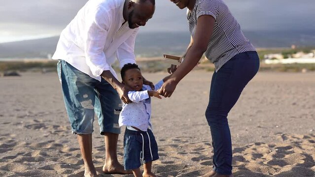 Happy african couple having playful time with toddler on the beach - Parents dancing with baby son outdoor