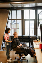 Vertical full length portrait of young woman with buzzcut sitting in beauty salon chair with female hairstylist working