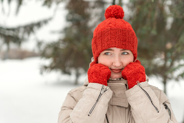 Cute girl in red knitted mittens and hat poses on winter forest or park background. Woman in comfortable winter clothes outdoors