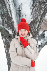 Smiling young woman in red hat and mittens stands near snow-covered tree. Portrait of cute thoughtfully girl looking to the side.