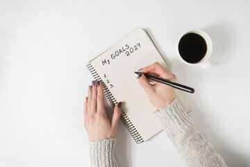 Female hands writing My Goals 2024 in a notebook. Mug of coffee on the table, top view