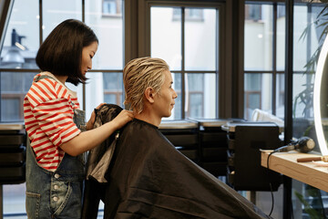 Obraz premium Side view portrait of young hairstylist putting protective cape on male client in beauty salon, copy space