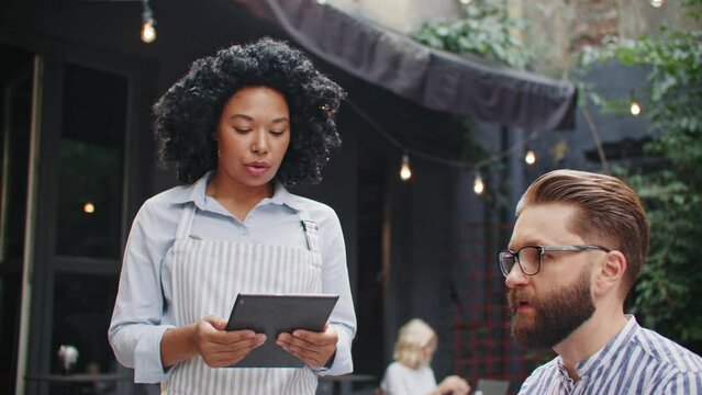 Handsome Caucasian man with beard ordering food at restaurant on terrace. African American waitress entering all of information into tablet device. Concept of modern world with digital technologies.