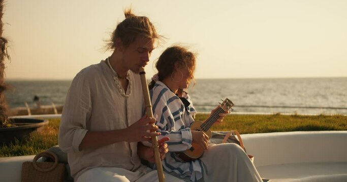 Happy musical duo guy plays the flute while the girl plays the ukulele near the sea on the beach. A guy plays the oboe on the beach near the sea. A blond guy in light clothes plays a wind musical