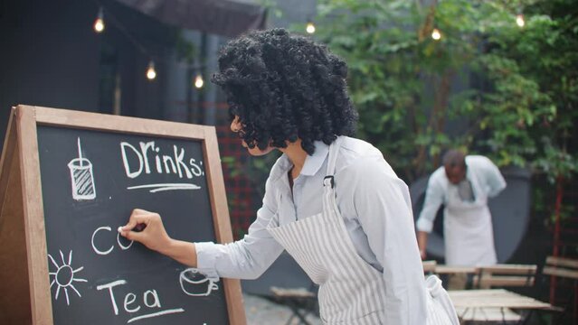 African American female owner writing on board menu of her small bakery shop. Happy woman owner wearing apron getting ready for receiving clients. In background male waiter cleaning tables on terrace.