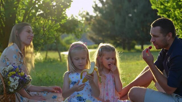 Close up of cute blonde little girls on family picnic with parents in summer. Caucasian children are eating fruits sitting on blanket. Happy childhood. Family weekend. Real time.