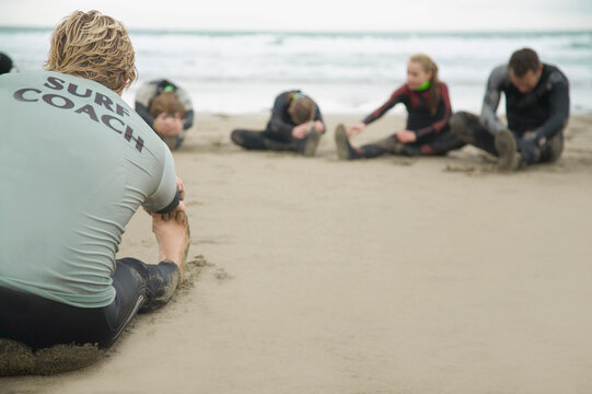 Back view of a surfing instructor and students stretching their legs on a beach
