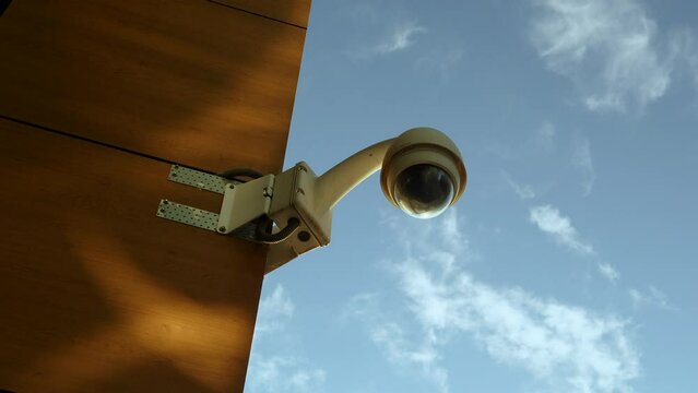 A security camera mounted on a rust-colored wall against a backdrop of blue sky.