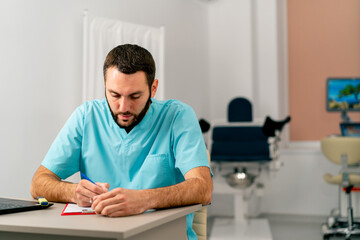 A male doctor in a medical uniform sits in medical office at his desk with a laptop and fills out papers - obrazy, fototapety, plakaty