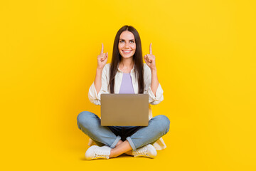 Full length photo of cheerful gorgeous person sit floor use netbook look direct fingers up empty space isolated on yellow color background