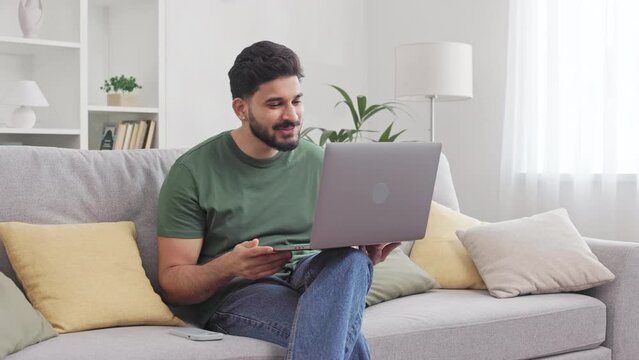 Smiling bearded man waving with hand for greeting interlocutor during video conference on modern laptop. Attractive indian male in casual clothes sitting on couch and enjoying online communication.