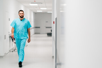 A tall male doctor with a beard walks along the hospital corridor with folder of documents and...