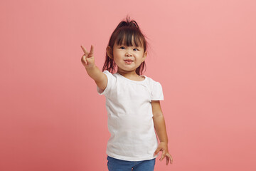beautiful ittle asian girl smiles and shows V gesture standing over pink isolated background.