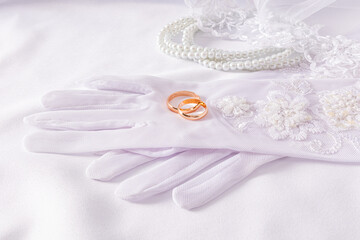 A pair of gold wedding rings lie on luxurious wedding white gloves. White satin background with pearl beads. Wedding accessories concept.