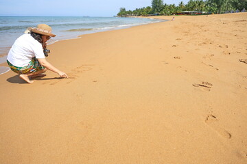 Female tourists drawing "LOVE" in the sand happily at the beach while visiting the sea. A woman sits with her back to the sea and is writing the letters to express the word LOVE on the fine sand. 
