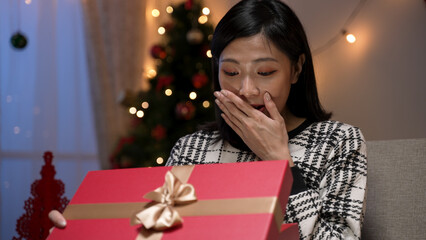 happy asian female covering her mouth with a surprised look on face as she opens and sees the beautiful Christmas present in a cozy living room at night